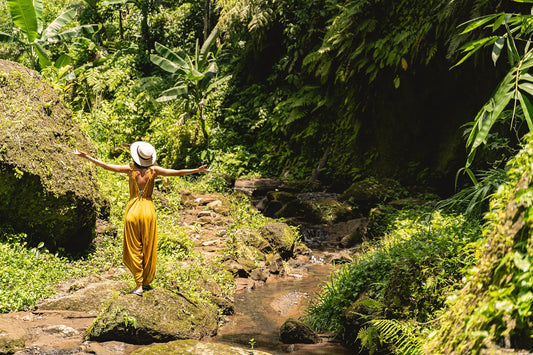 A woman in a flowing mustard yellow jumpsuit and white straw hat stands on mossy rocks beside a stream in nature