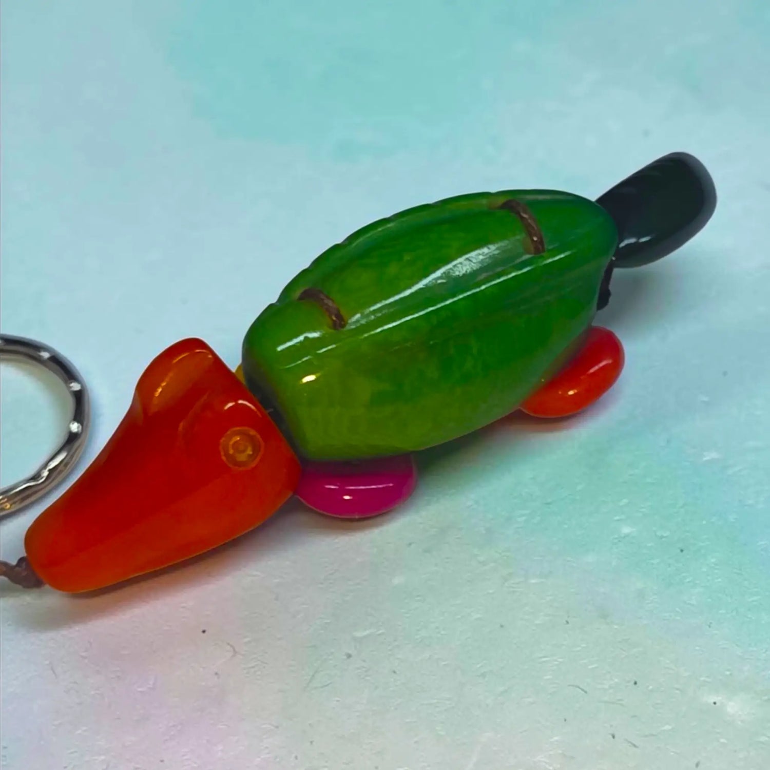 A vibrant hand-carved animal keychain platypus with green body orange head and feet pink underbelly and metallic ring attachment