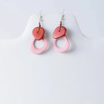 A pair of charli geometric tagua earrings with coral red heart tops and soft pink oval rings connected by brown cord