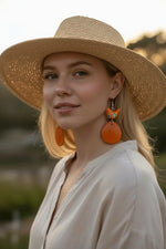 A wide-brimmed natural beige straw hat with woven texture featured alongside folk art orange bird tagua slice earrings