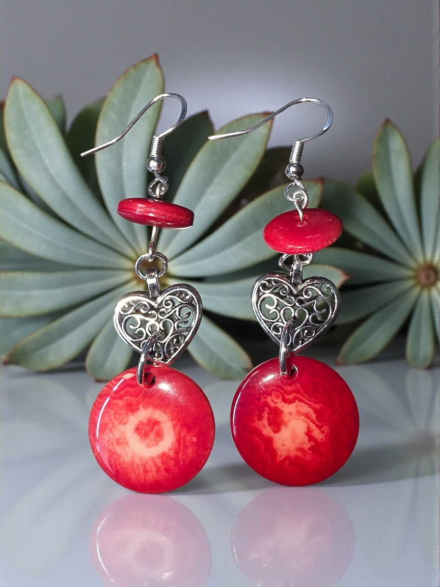 Red earrings with silver heart details on a reflective surface with succulent plants in the background