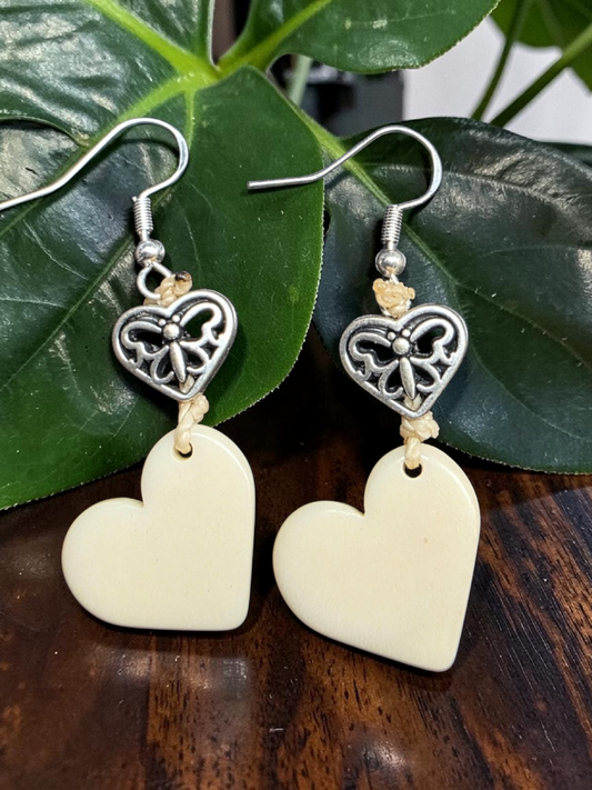 Heart-shaped carved tagua earrings with silver filigree design on a wooden surface with leaves in the background