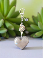 A silver-toned keychain with a glossy gray ceramic heart, ammonite shell charm, and clear crystal bead bag charm
