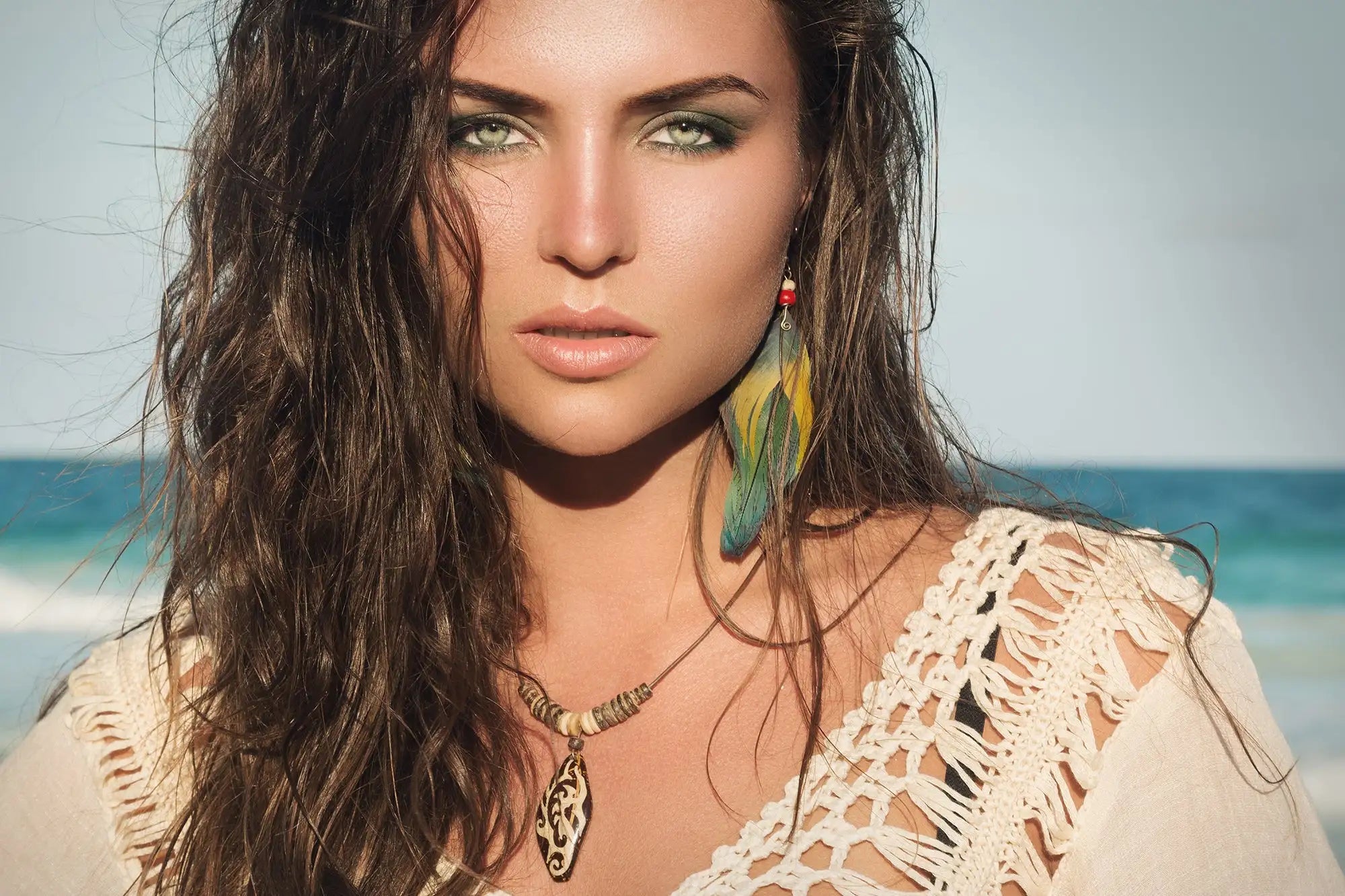 A woman wearing a cream-colored crochet top with intricate lace detailing and layered necklaces, including one featuring an ornate pendant.
