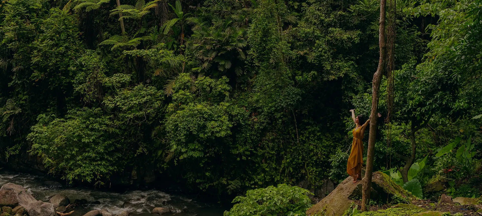 A woman in a flowing golden-orange dress stands beside a tall tree, surrounded by lush green jungle foliage.