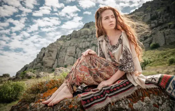 A woman with long, flowing red hair sits barefoot on a mossy rock wearing an earth-toned patterned dress over a light beige blouse.