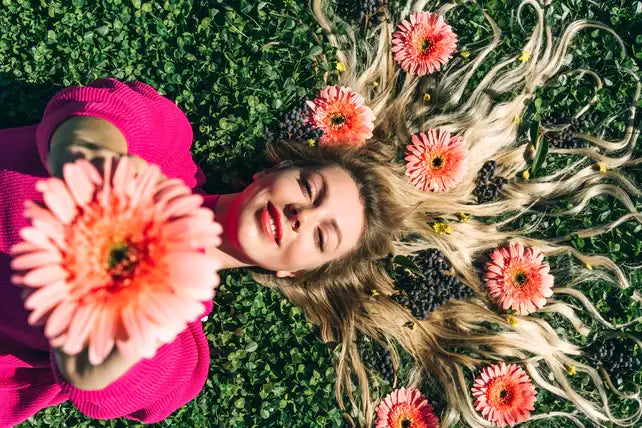 A woman in a vibrant pink sweater lies on green grass, holding a large peach-colored gerbera daisy close to her face.