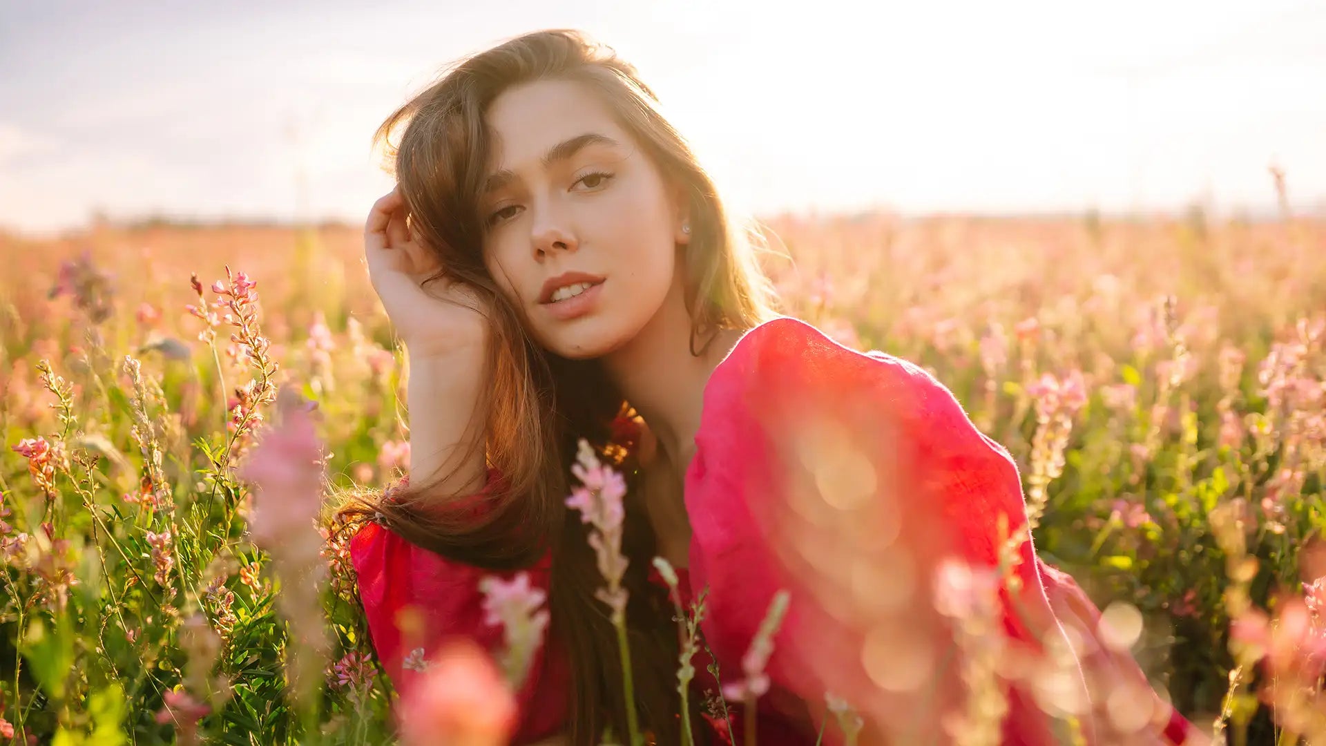 A woman in a vibrant red, flowing garment stands amidst tall pink and green wildflowers under warm sunlight.